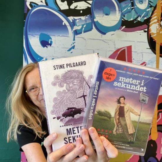 Lejla står op af en farverig væg. Hun holder "Meter i sekundet" som bog og film i hænderne foran sit ansigt..