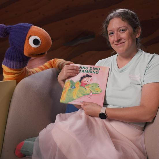 En perso sidder i en blød stol og viser bogen Fino Dino Bambino frem. Til venstre for sidder en stor dukkeudgave af Orm fra DR Minisjangs Børsteuniver. Billedet stammer fra "Læs med Orm"