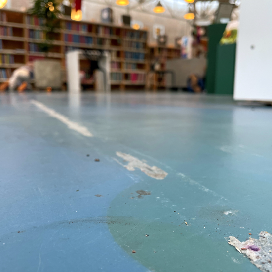 Et halvklat og slidt linoliumsgulv i frøperspektiv. I baggrunden aner man bogreoler og børn på biblioteket.