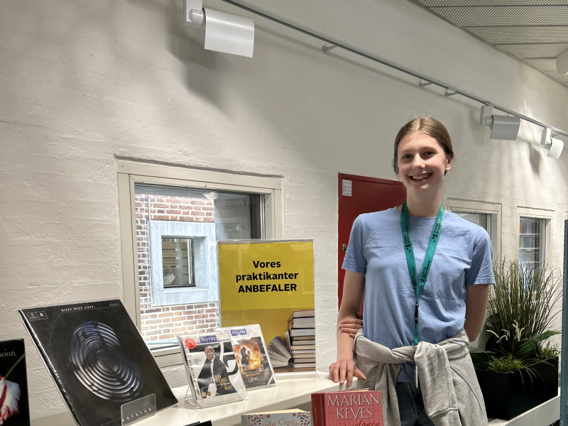 Frederikke står ved siden af sin udstilling på Hovedbiblioteket. På udstillingen ses forskellige bøger, film og musik. Frederikke smiler til kameraet.