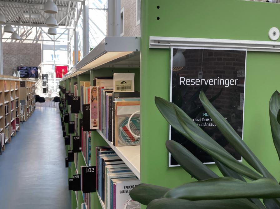 De grønne reserveringshylder på Hovedbiblioteket. I forgrunden bag en plante står et skilt med "Rerserveringer" skrevet på.